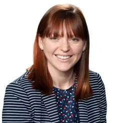 A headshot of Carolyn Nikoloski, a smiling female with red hair wearing a striped navy jacket