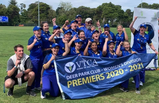 Group photo of Deer Park women's division cricket club holding a large, triangular premiers flag. The flag says: "VTCA Deer Park Club - Women's Division Premiers 2020-2021"